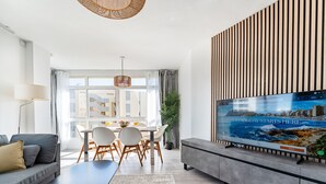 Ein modernes Wohnzimmer mit Essbereich, einem großen Fernseher, der ein Urlaubsziel zeigt, und Blick auf die Stadt durch große Fenster.