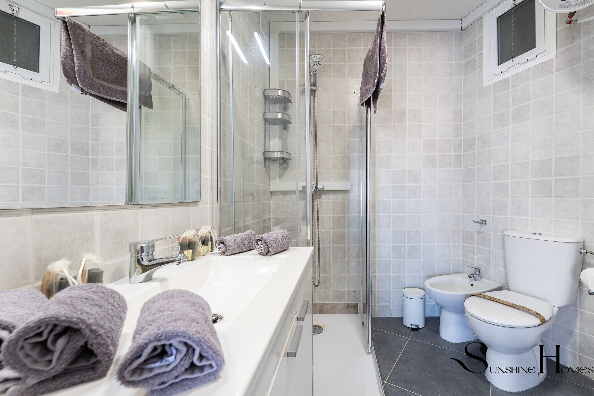 Moderni kylpyhuone, jossa on lasiseinällä varustettu suihku, valkoinen pesuallas peilillä sekä kaksi wc-istuinta.