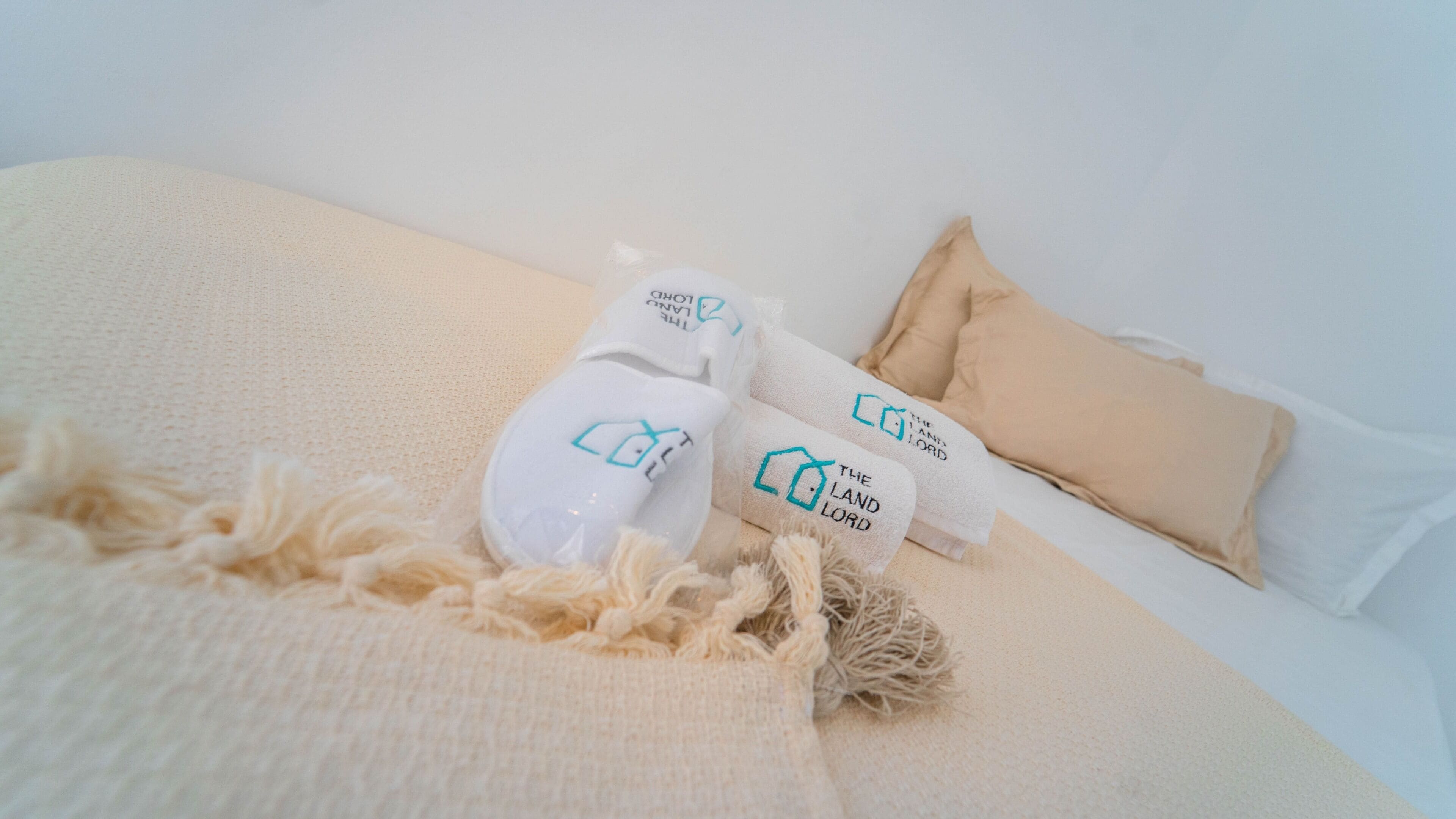 Ein Bett mit einer beigen Decke, zwei weiße Slipper mit blauer Aufschrift und ein beiges Kopfkissen.