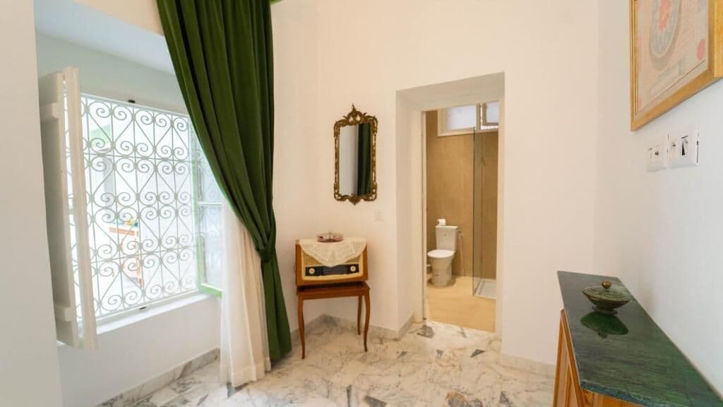 Une salle de bain avec un sol en marbre, une chaise en bois, un miroir, des toilettes et un meuble vert.