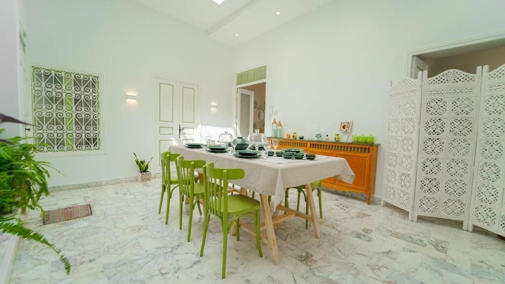 Un comedor con suelo de mármol, sillas verdes, una mesa dispuesta con platos y vasos, y una partición blanca decorativa.
