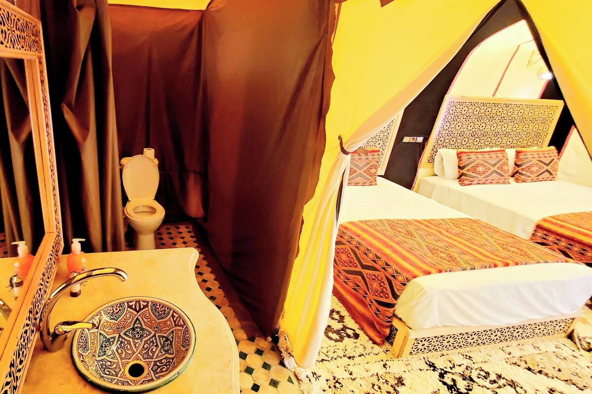 Une pièce comprenant un grand miroir décoratif, un lavabo orné de carreaux, des toilettes et deux lits avec une literie à motifs.