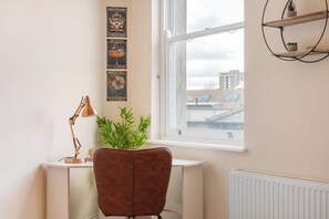 A room with a desk, a chair, a potted plant, a radiator, and a window with a view of buildings. - Bright 1BR Apartment in Shoreditch Brick Lane Close to Tube Sleeps 4 (London)
