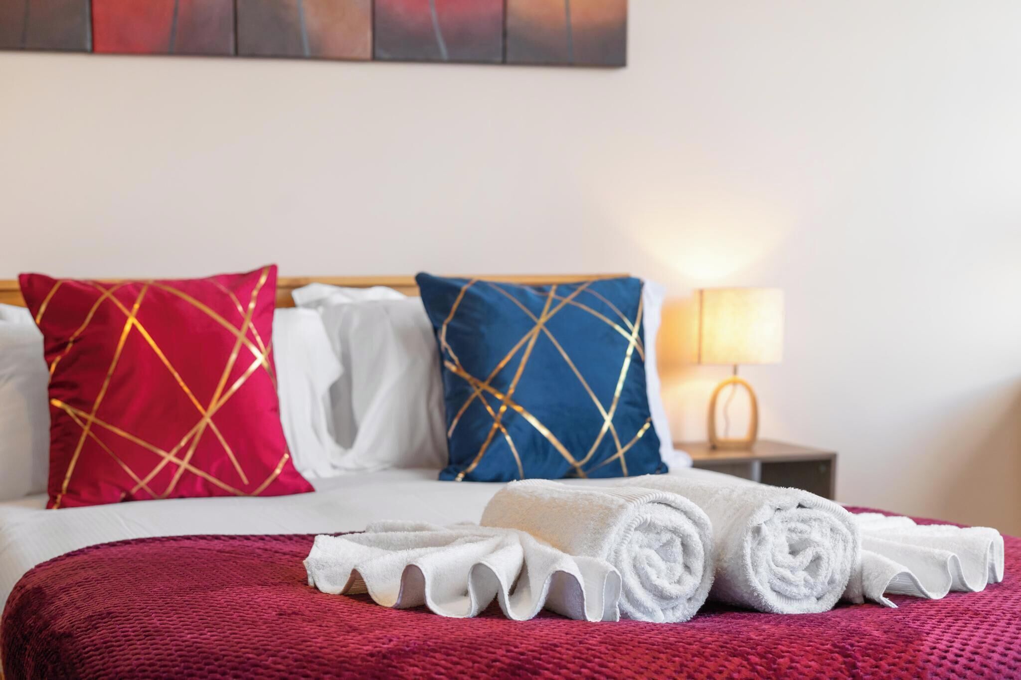 A hotel room with a bed, pillows, a red blanket, and rolled towels.