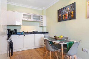 A compact kitchen with white cabinets, a black countertop, and a dining area with a table and chairs. - Bright 1BR Apartment in Shoreditch Brick Lane Close to Tube Sleeps 4 (London)