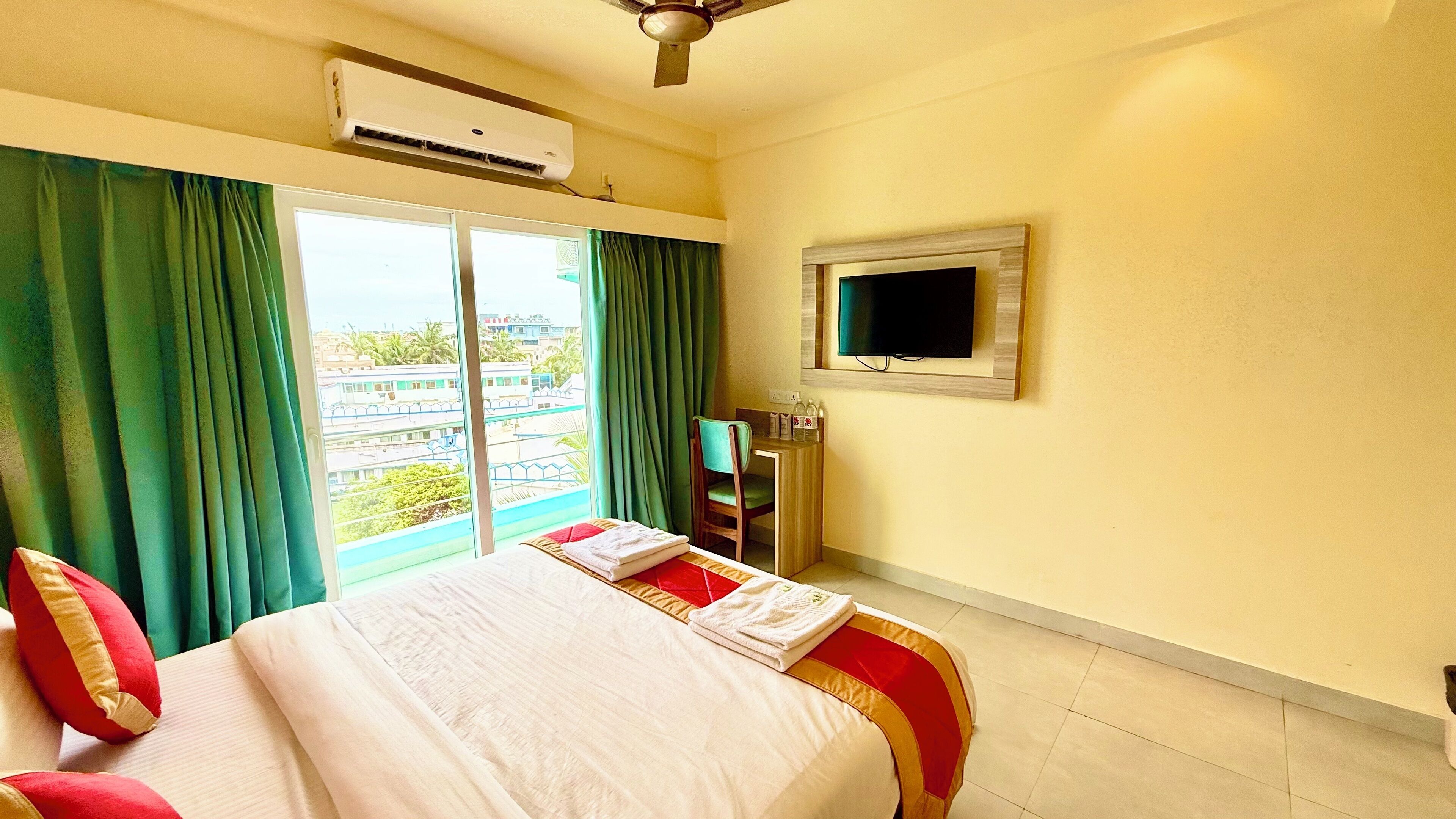 A hotel room with a bed, green curtains, a flat-screen TV, and an air conditioning unit.
