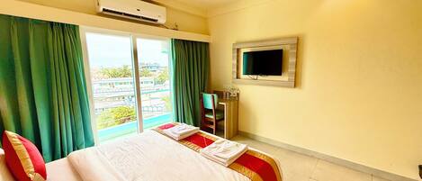 A hotel room with a bed, green curtains, a flat-screen TV, and an air conditioning unit.