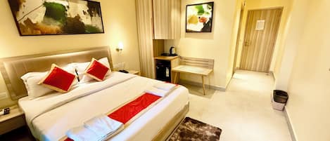 A hotel room with a large bed, a desk, a chair, a small cabinet, and a trash bin.