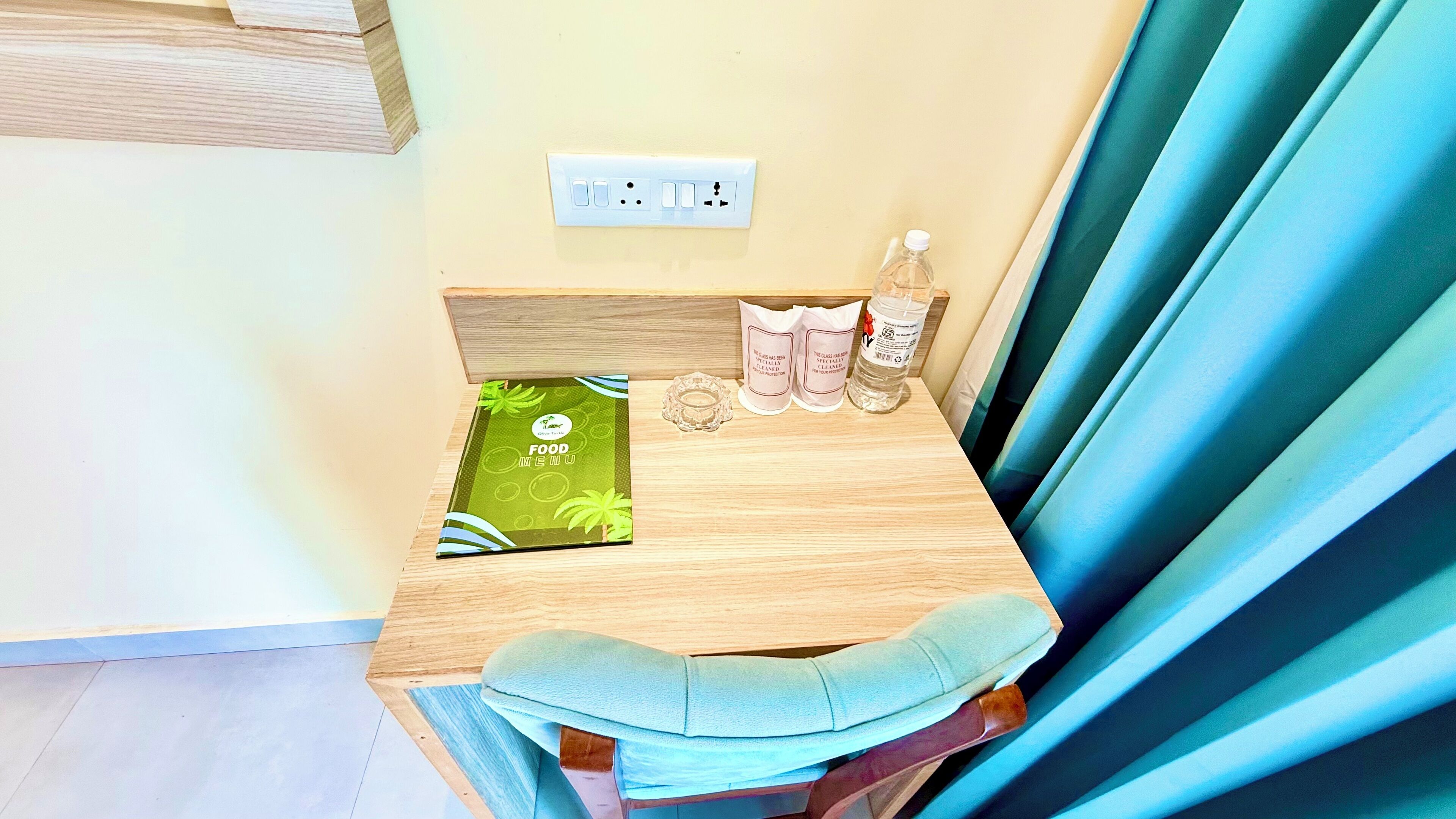 Une petite table en bois, un menu vert, une bougie dans un verre, une bouteille d’eau et une paire de chaussons.