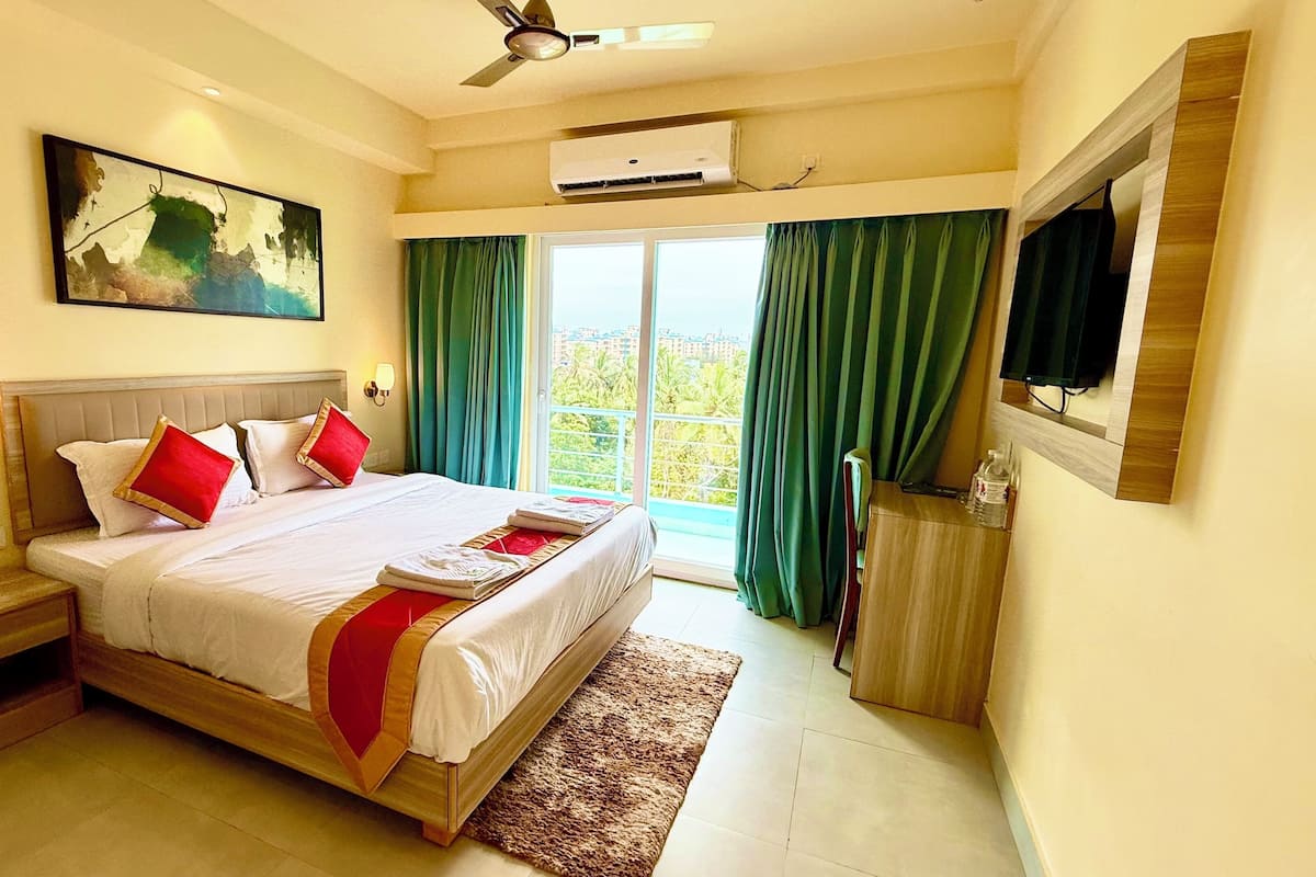 Una habitación de hotel con cama, mesita de noche, lámpara de noche, televisor montado en la pared, equipo de aire acondicionado y ventana con cortinas.