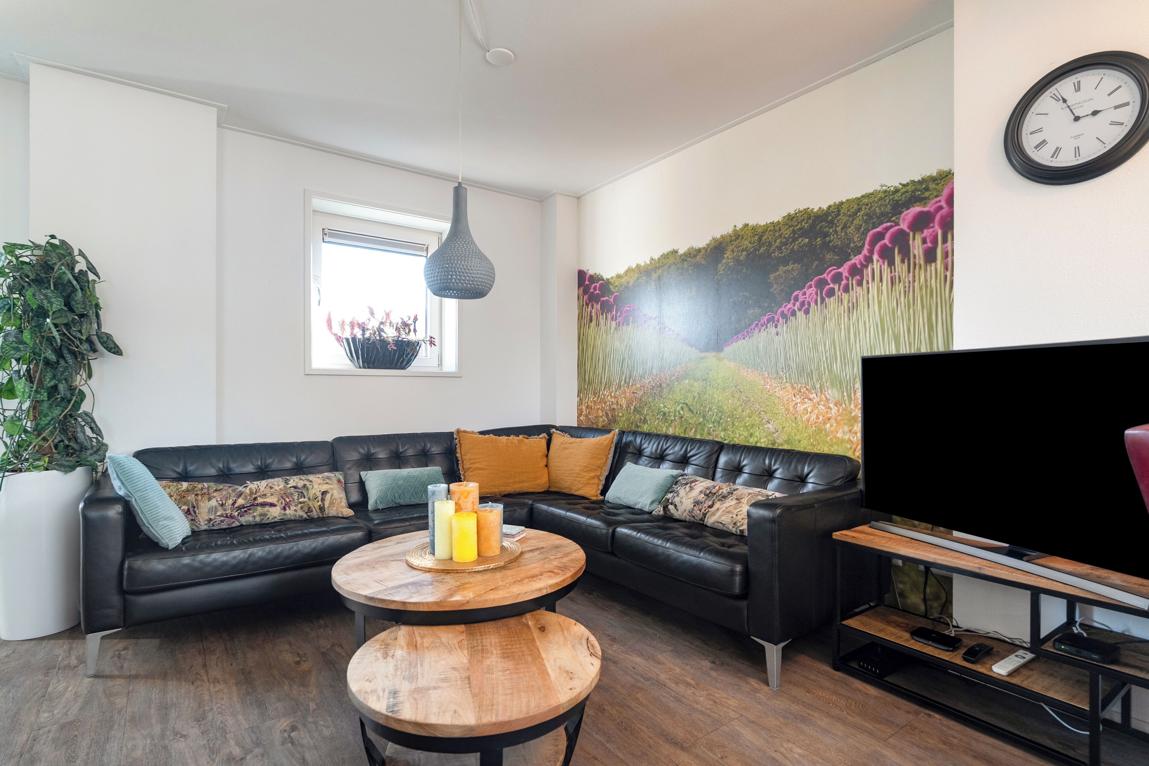 A modern living room with a black leather sofa, a round wooden coffee table, a large flat-screen TV, and a wall-mounted clock.