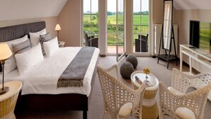 Habitación de hotel con cama, escritorio, televisión, sillas de mimbre y un balcón con vistas al campo.
