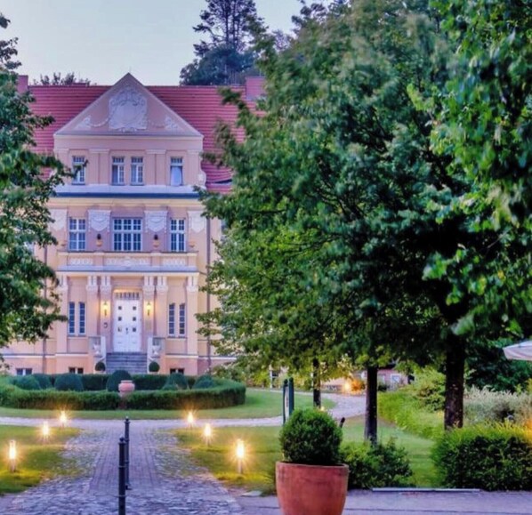 Un grand bâtiment imposant, doté d’une façade symétrique, d’une entrée centrale et d’un jardin bien entretenu, avec un chemin pavé menant à l’entrée.
