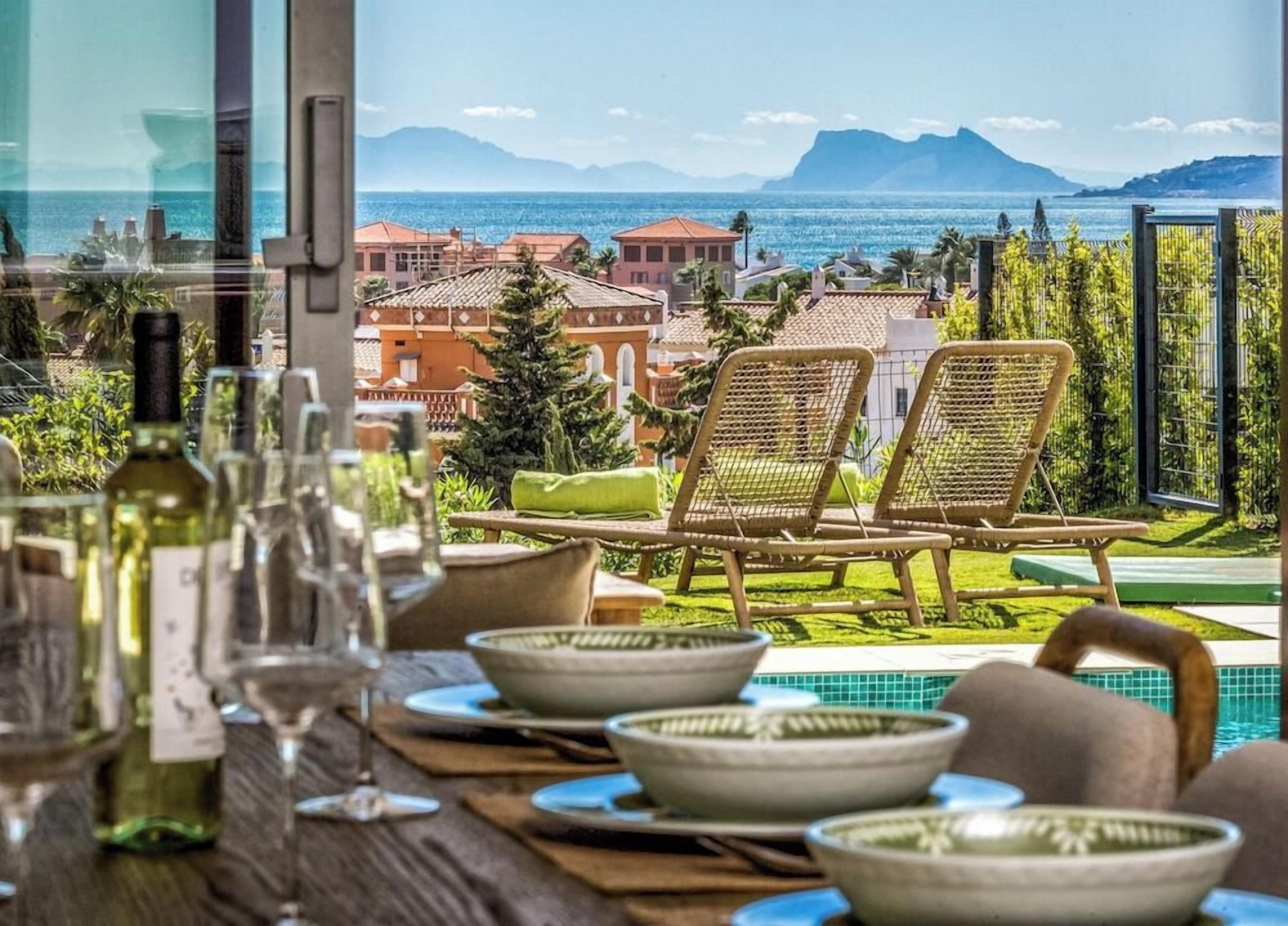 A dining area with a view of the sea and mountains, featuring wicker chairs and a poolside setting.