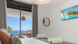 Ett modernt hotellrum med en balkong, ett stort fönster med utsikt över havet, en platt-TV på väggen och två handdukar med hotellets logotyp.