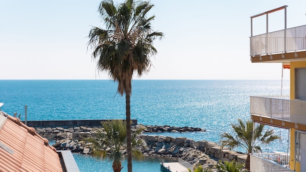 Vista de una zona de piscina con una palmera, una costa rocosa y un edificio con balcón.