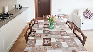 A dining table with a floral tablecloth, surrounded by chairs, in a kitchen with white cabinets and a wooden door. - Relaxation in Holiday House Calmasino (Veneto)