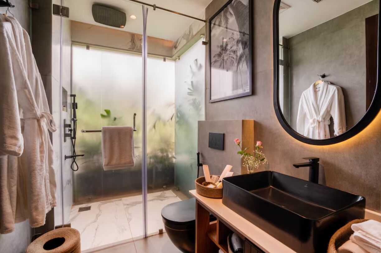 Un baño moderno con cabina de ducha de vidrio, un lavamanos tipo cuenco negro y un espejo redondo.