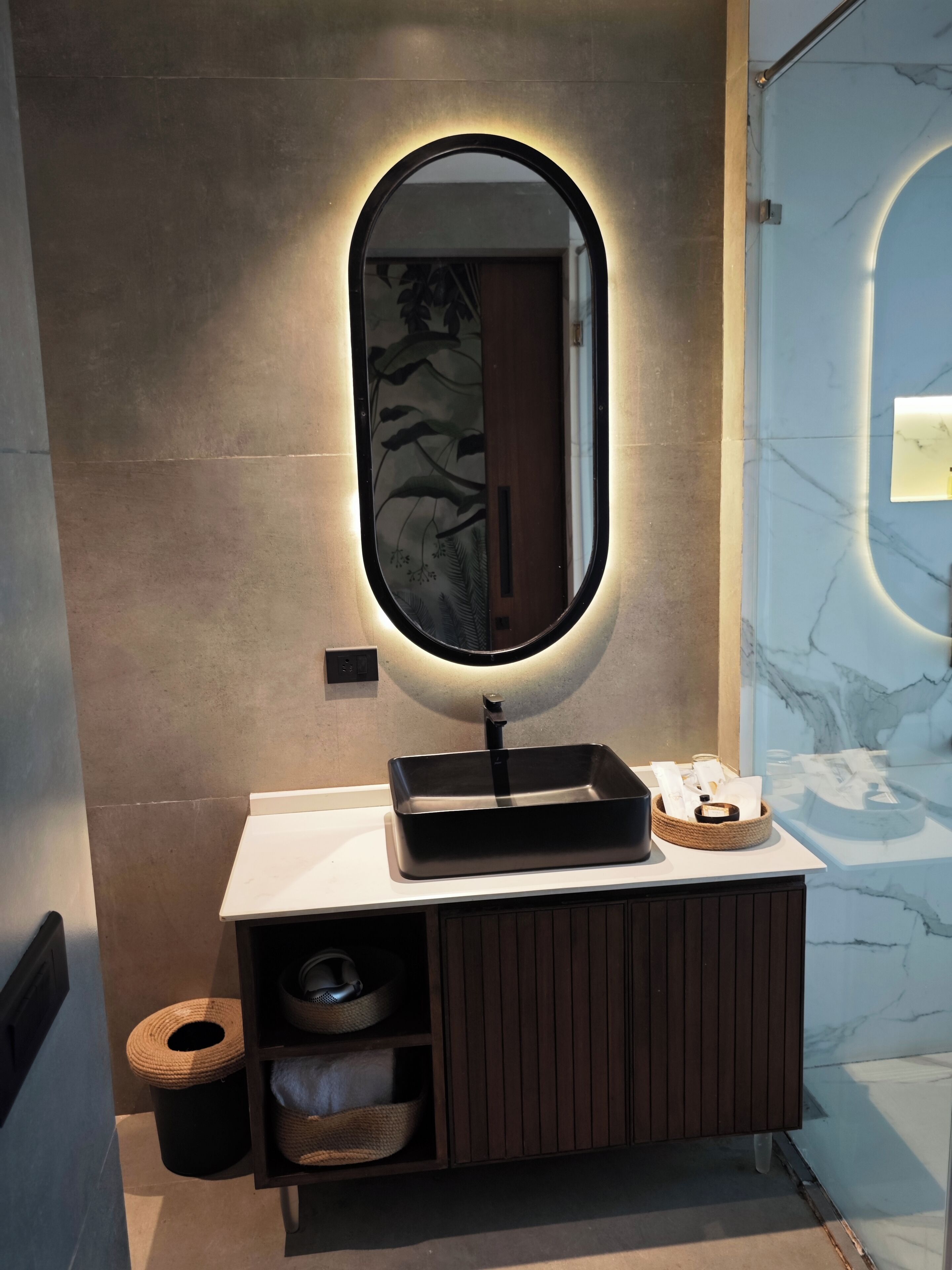 A bathroom with a modern vanity, a large oval mirror with underglow lighting, a marble wall, and a wooden cabinet with storage baskets.