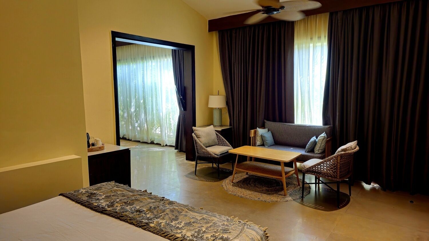 A hotel room with a bed, a sofa, two chairs, a small table, a lamp, a ceiling fan, and a view through a window with curtains.