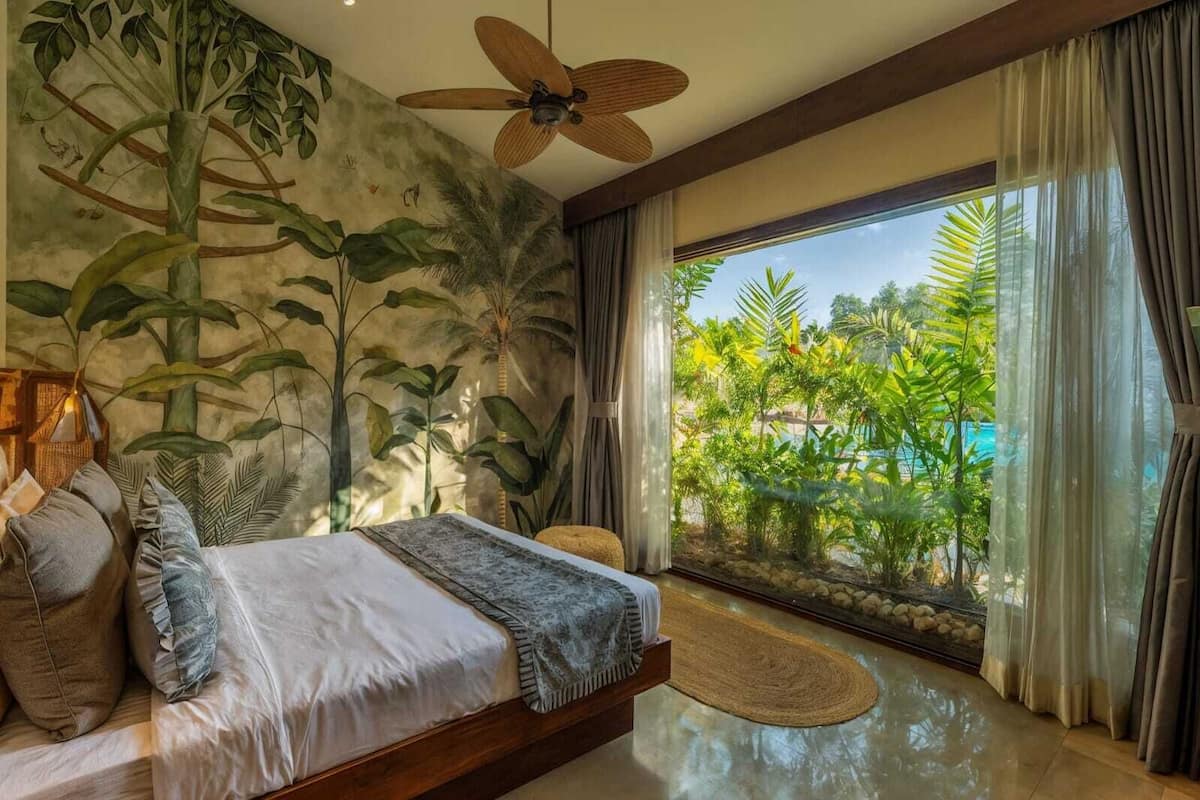 Un dormitorio con una cama grande, un ventilador de techo y vistas a un jardín tropical a través de la ventana.