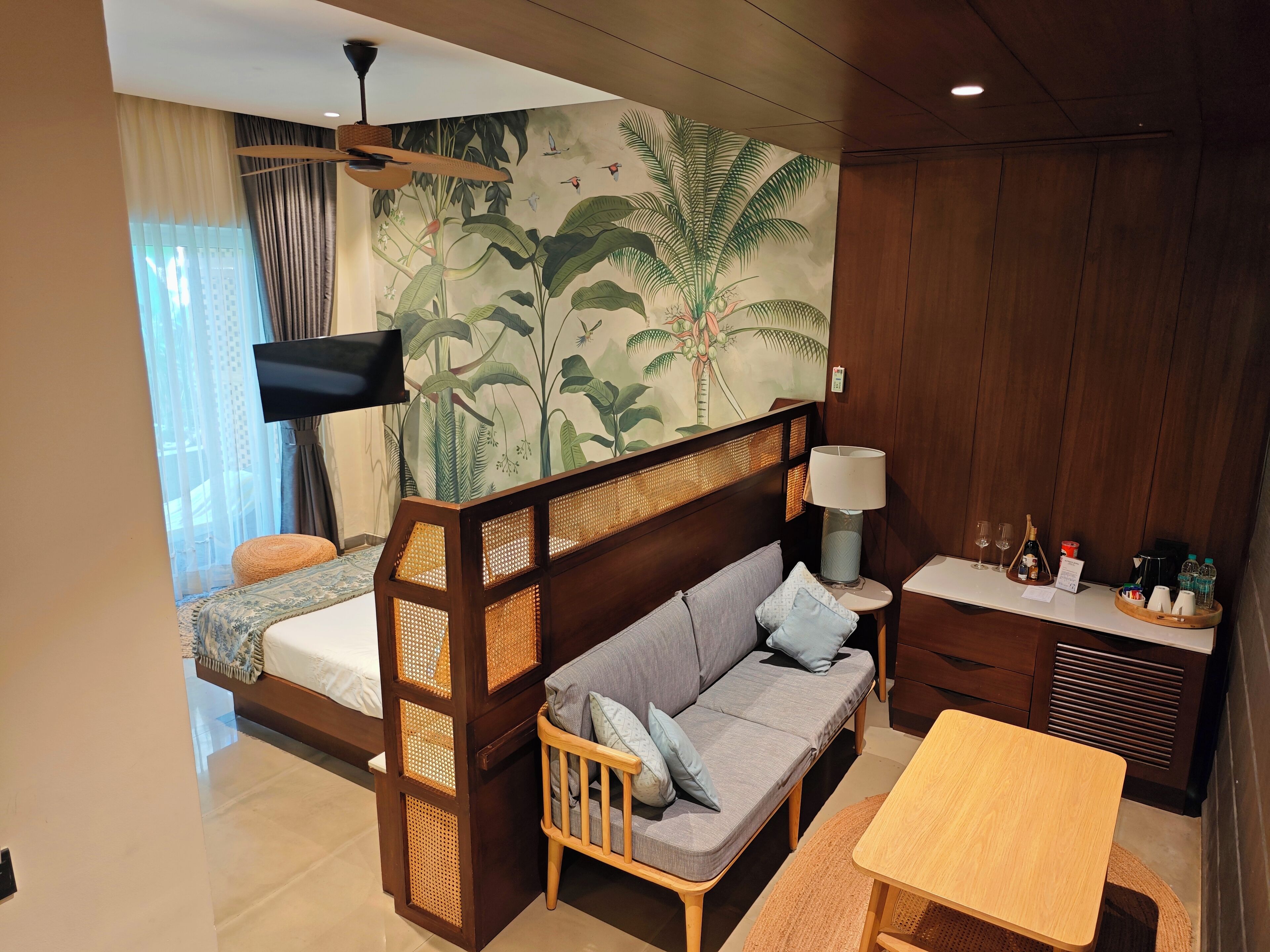 Una habitación con cama, sofá, televisor, mesa y pared decorada con un mural tropical.