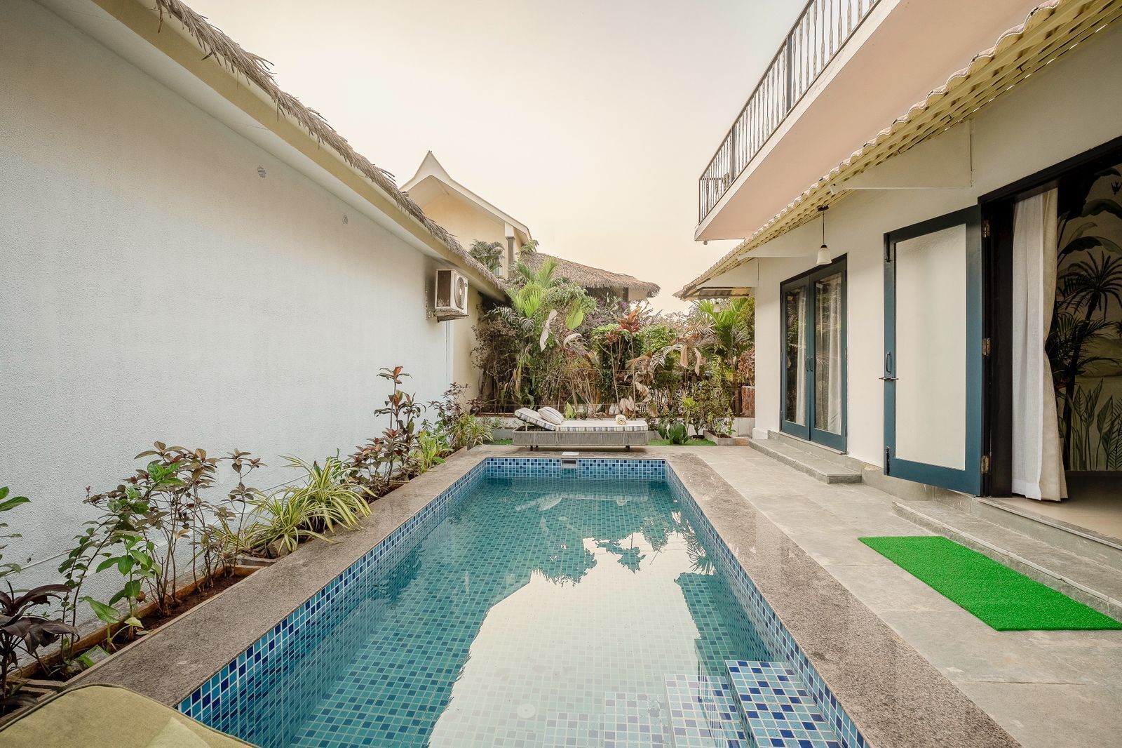 A swimming pool with tiled edges, surrounded by plants and a small seating area.