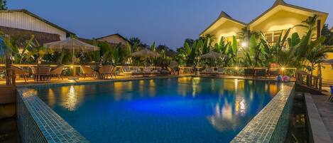 A swimming pool with lounge chairs and umbrellas, surrounded by buildings and palm trees.