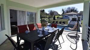 Eine Außenpatio mit einem Esstisch, gedeckt für vier Personen, umgeben von roten Stühlen. Im Hintergrund ist ein Pool mit einem Liegestuhl und ein kleiner Garten zu sehen.