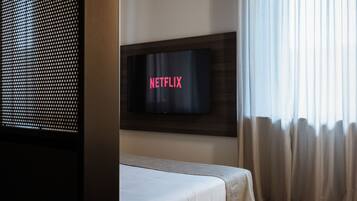 Ein Schlafzimmer mit einem Bett, einem an der Wand montierten Fernseher und einem Fenster mit Vorhängen.