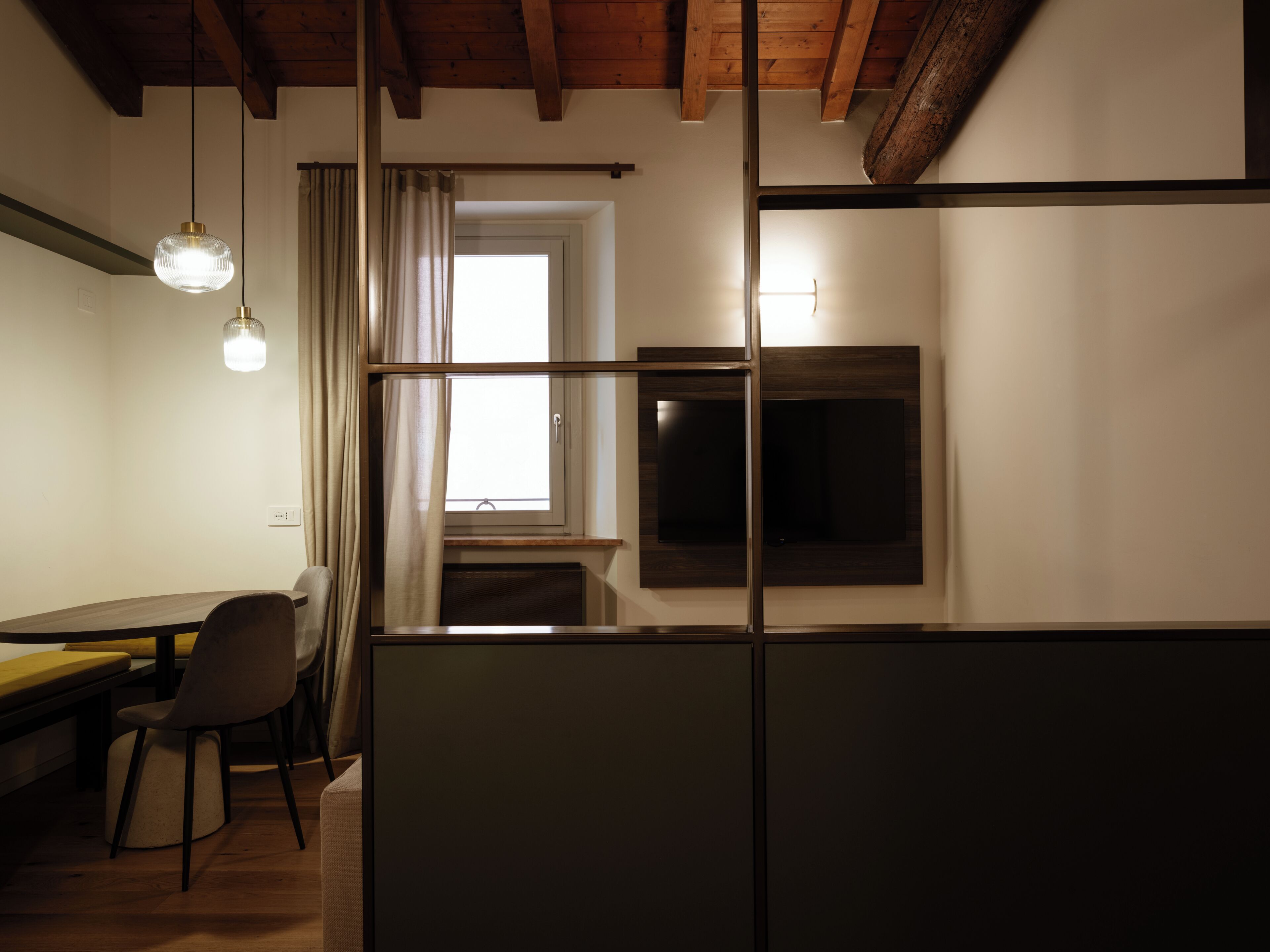 A room with a wooden ceiling, a dining table with chairs, a TV mounted on the wall, and a window with curtains.