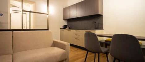 A modern kitchen with wooden cabinets, a black countertop, and a built-in stove. There is a beige sofa and a gray chair in the living area.