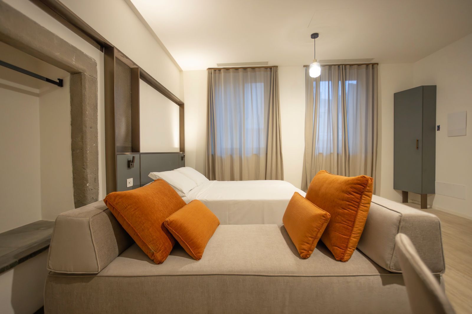 Quarto de hotel com cama, sofá com almofadas laranja, guarda-roupa e janela com cortinas.