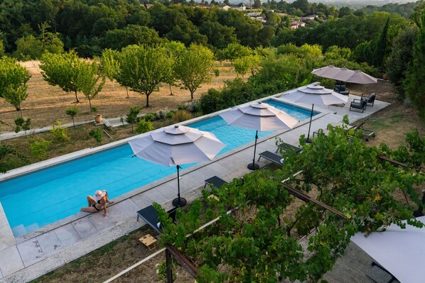 Una piscina con sillas de descanso y sombrillas, rodeada de árboles y un viñedo.