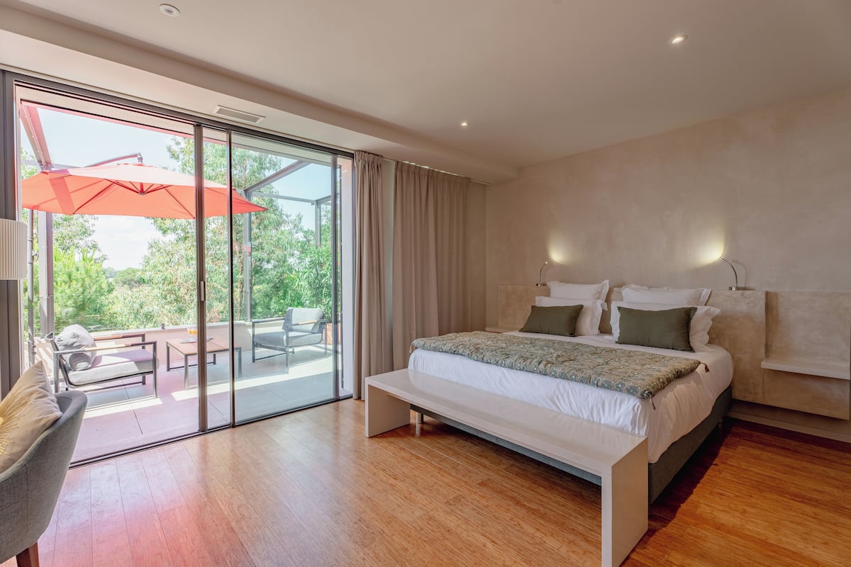 A modern bedroom with a large bed, a bench, a chair, and a sliding glass door leading to an outdoor area with a red umbrella.