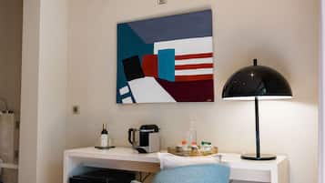 A hotel room with a desk, a coffee maker, a lamp, and a painting on the wall.