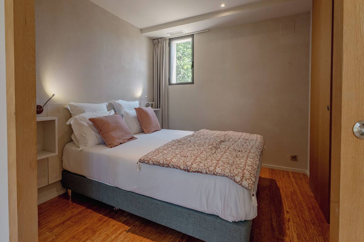 A bedroom with a bed, pillows, a bedside table, a lamp, a window, and a door.