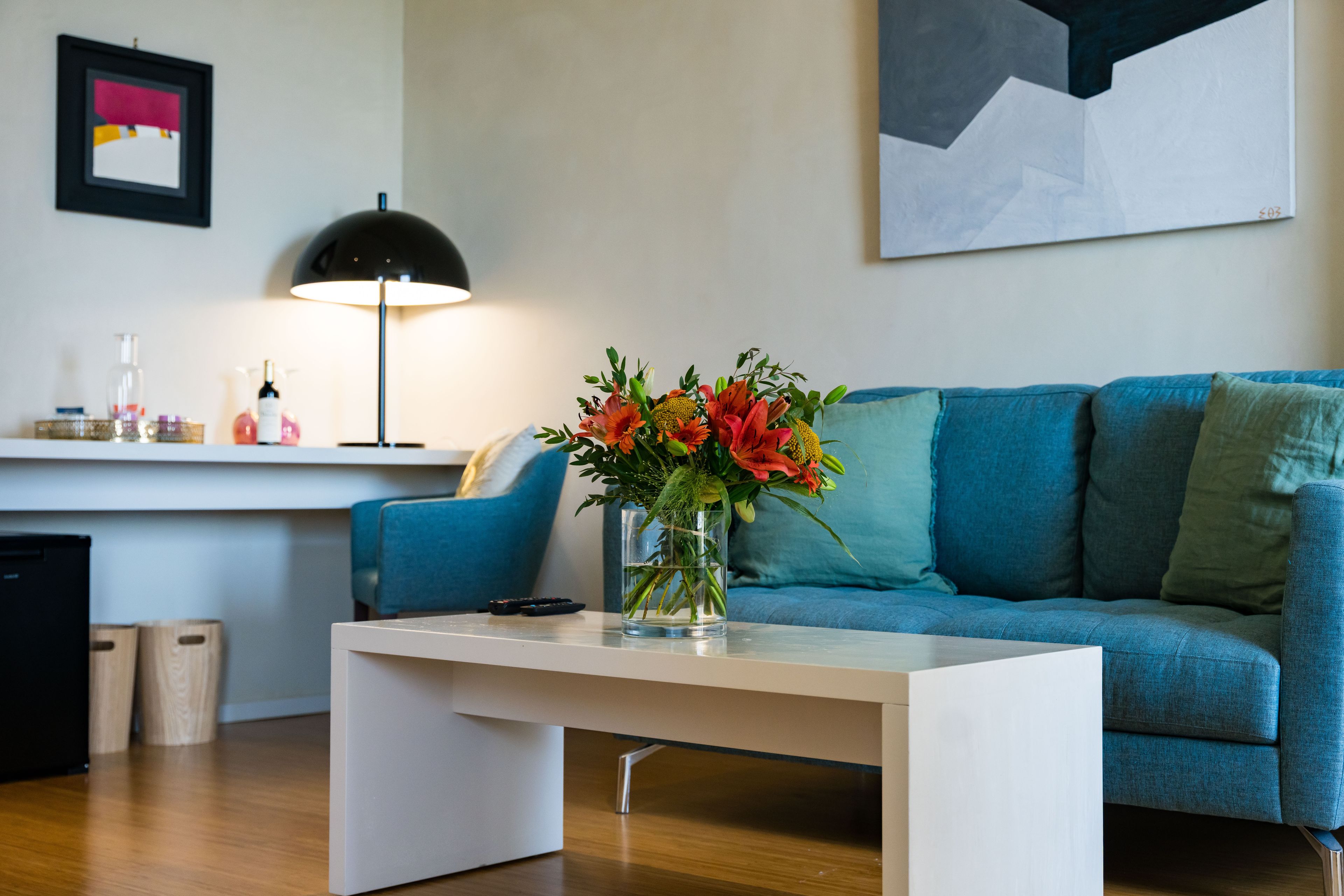 Una sala de estar moderna con un sofá azul, una mesa de centro blanca con un florero y una lámpara de pie negra, además de un cuadro en la pared.