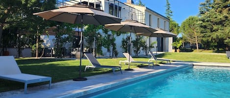 Piscina com espreguiçadeiras e guarda-sóis, rodeada por um gramado bem cuidado e um edifício ao fundo.