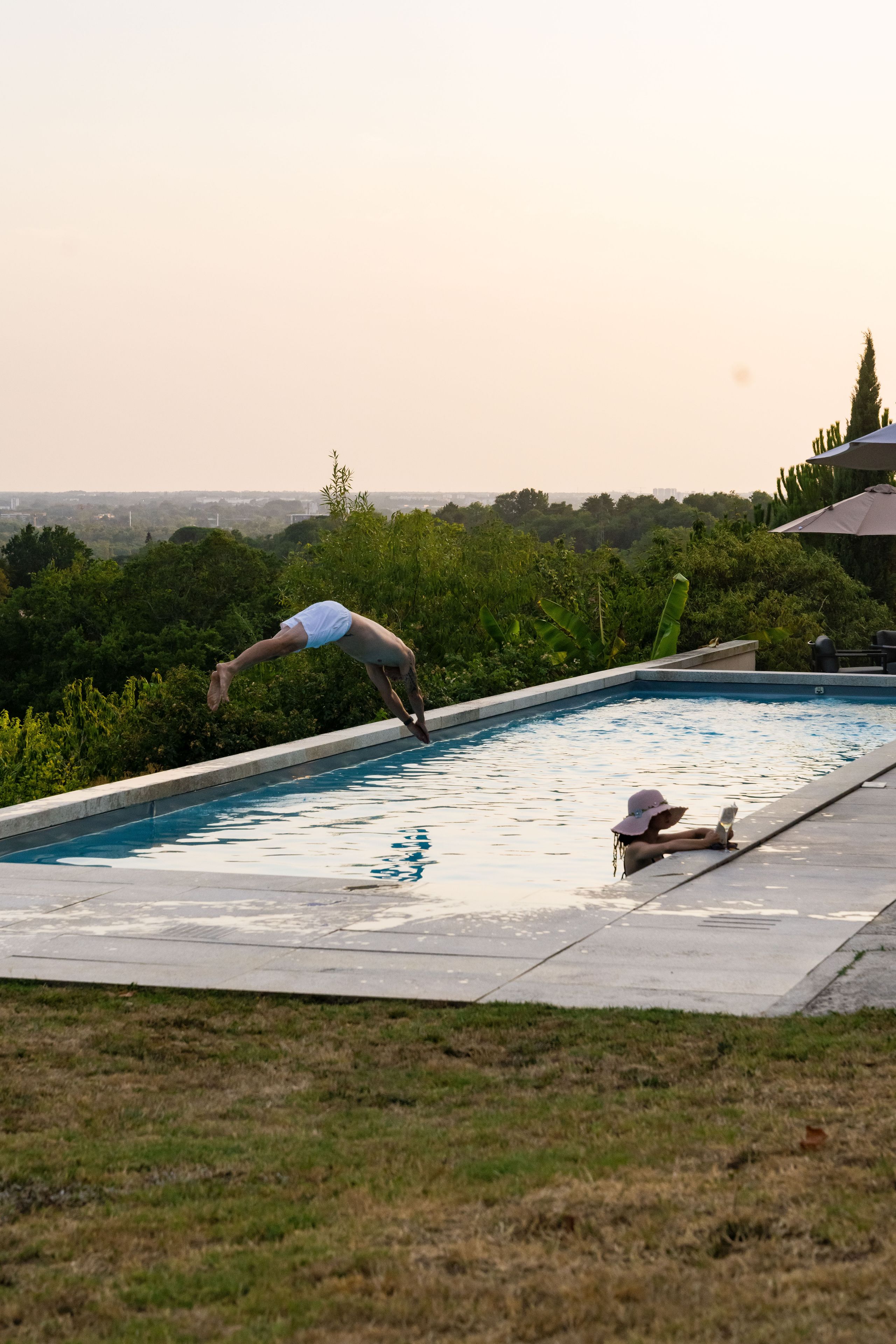 A swimming pool with a person diving into it, surrounded by greenery and a clear sky.