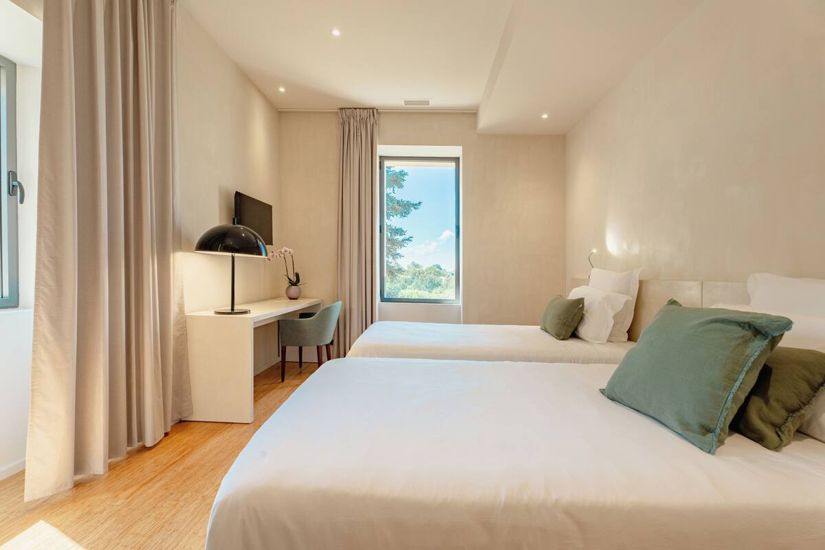 A modern hotel room with two beds, a desk, and a large window.