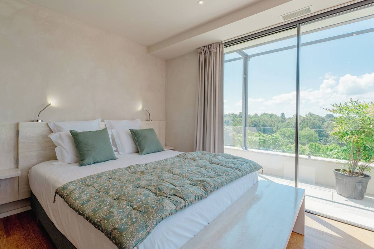 A modern bedroom with a large bed, a view of greenery outside, and a potted plant on the windowsill.