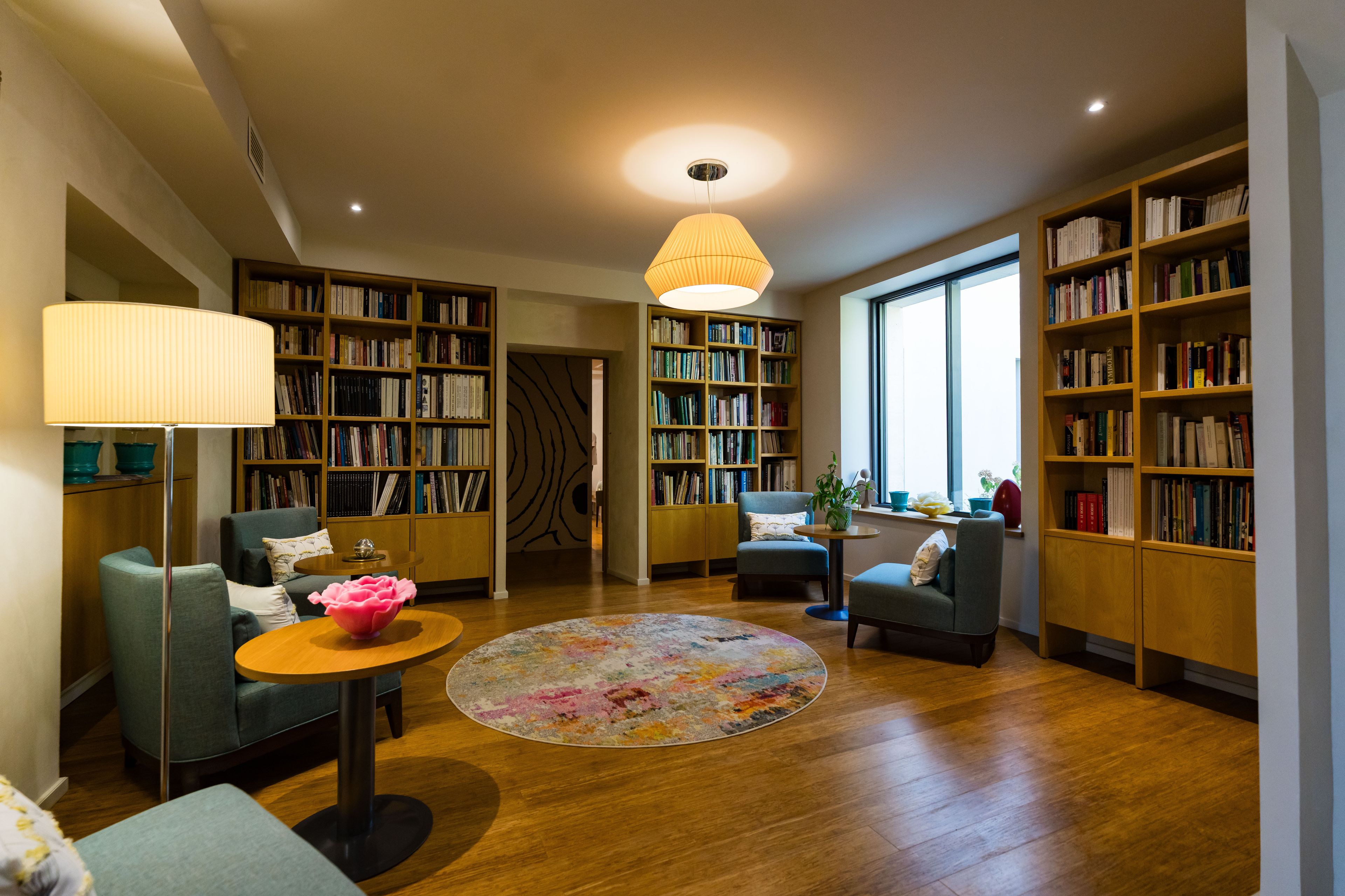 A spacious reading room with multiple bookshelves, comfortable seating, and a large window providing natural light.