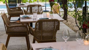 An outdoor dining area with wicker chairs and tables, set under a canopy, overlooking a body of water.