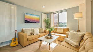 A living room with a sofa set, a coffee table, a guitar, and a large window with a view.