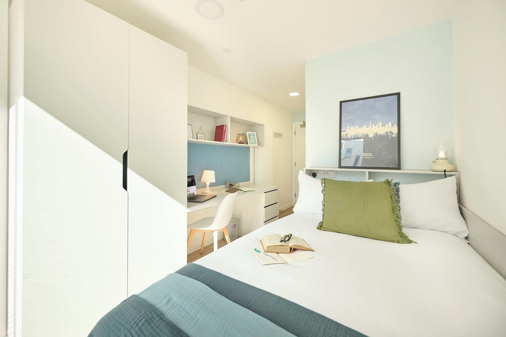 Une chambre à coucher avec un lit, un bureau et une chaise. Il y a un tableau encadré au mur et une lampe sur le bureau.