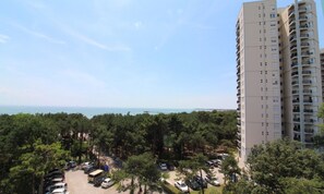 A high-rise building with a parking lot in front, surrounded by trees and a view of the sea. - Bright sea view apartment (Lignano Sabbiadoro)