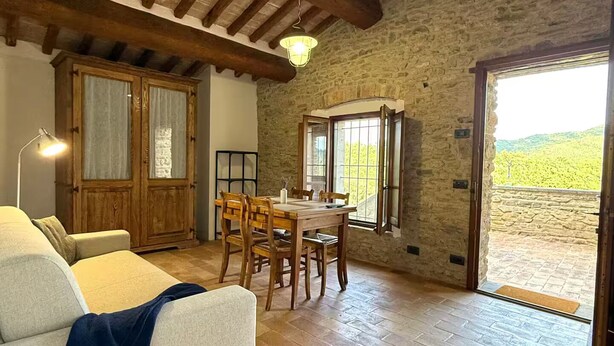 Ein gemütliches Wohnzimmer mit einer Couch, einem Holzesstisch und Stühlen. Man sieht eine Steinwand und Holzbalken.