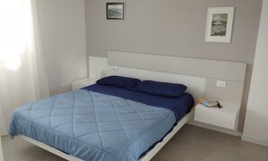A single-bed room with a white headboard, a blue quilted bedspread, and two pillows. There are two framed pictures on the wall, a white nightstand with a book and a phone, and a window with curtains. - Flat on the shores of Lignano (Lignano Sabbiadoro)