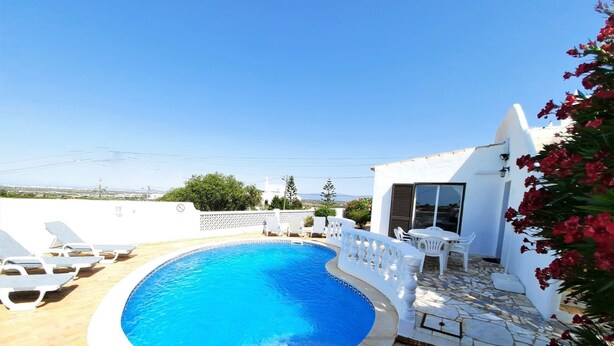 A swimming pool with lounge chairs, a white building with a tiled patio, and a view of the sea.